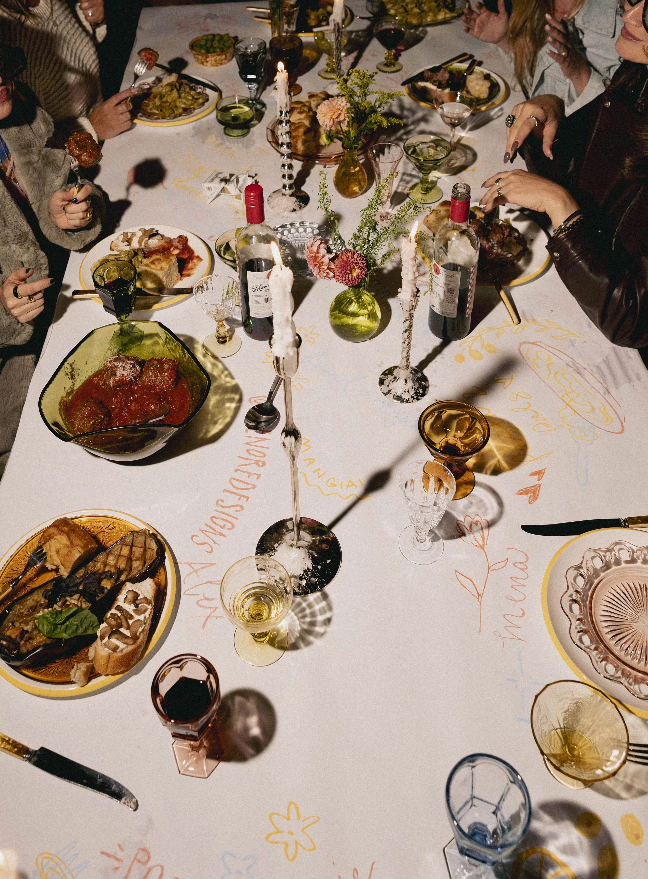 A festive dinner table with various dishes, glasses of wine, a vase with flowers, candles, and people enjoying a meal.