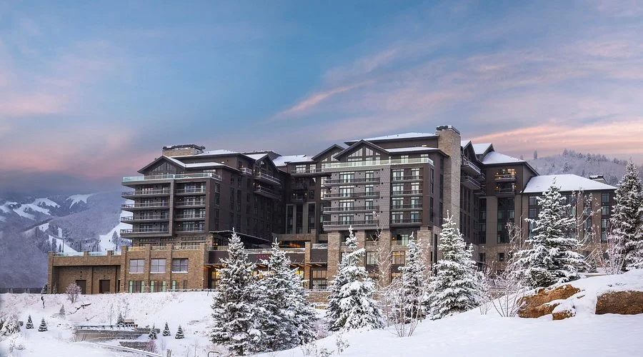 A modern multi-story hotel or apartment building in a snowy mountain landscape with pine trees in the foreground, under a colorful sunset sky.