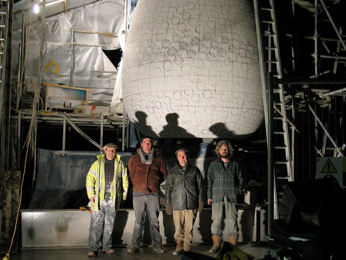 Four men standing in front of a large industrial construction site with a giant spherical structure overhead, casting their shadows on it.