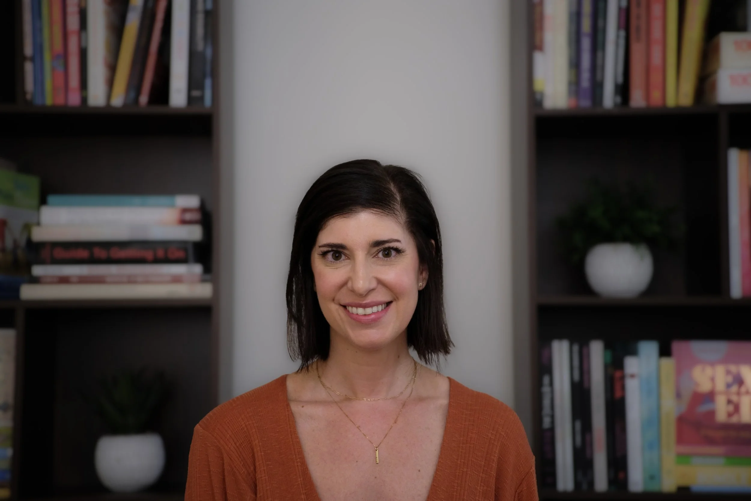 A woman with shoulder-length dark hair, smiling, wearing an orange top and a delicate gold necklace, standing in front of bookshelves filled with colorful books and houseplants.