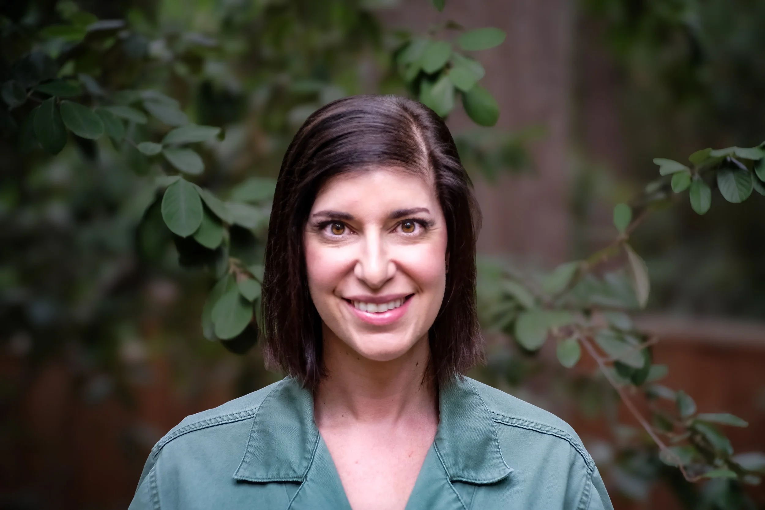 A woman smiling outdoors, with greenery and leaves in the background.