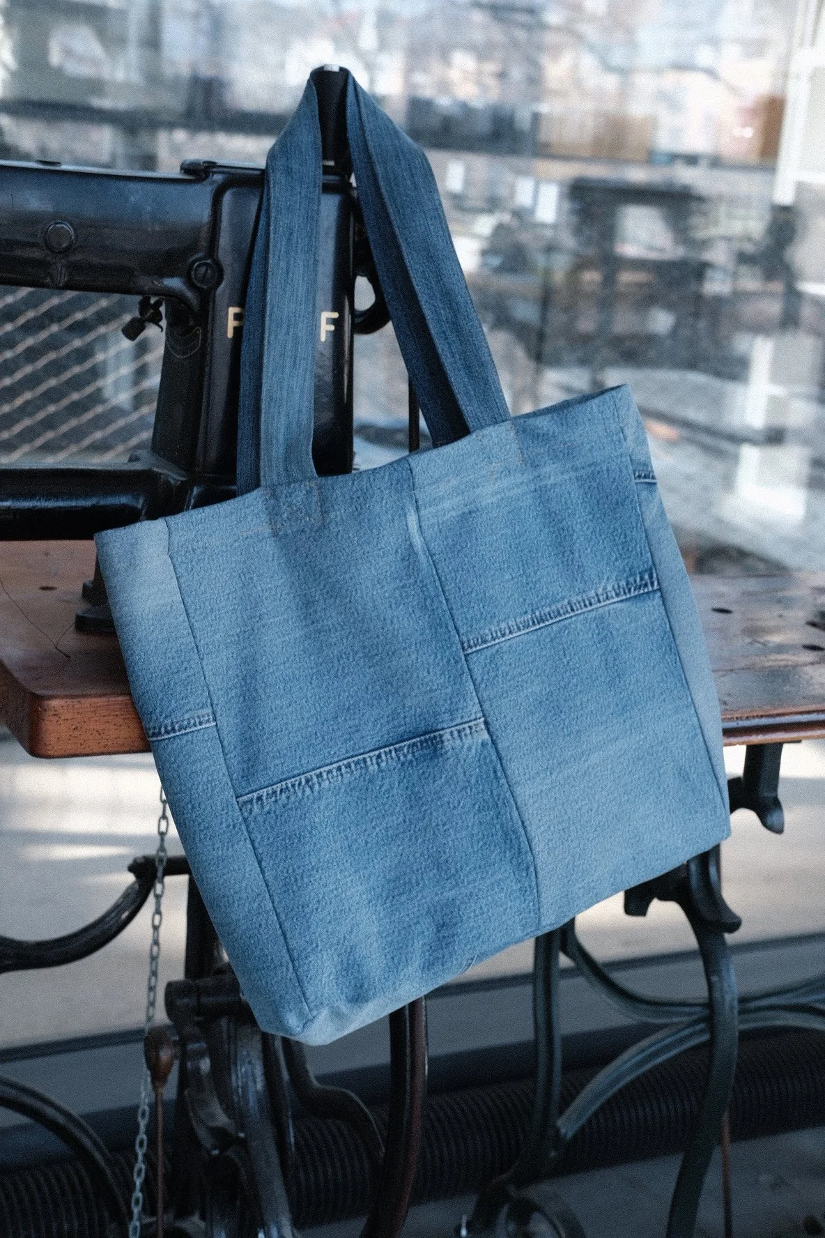 A blue denim tote bag hanging on a black metal chair backrest next to a vintage sewing machine on a wooden table with a window in the background.