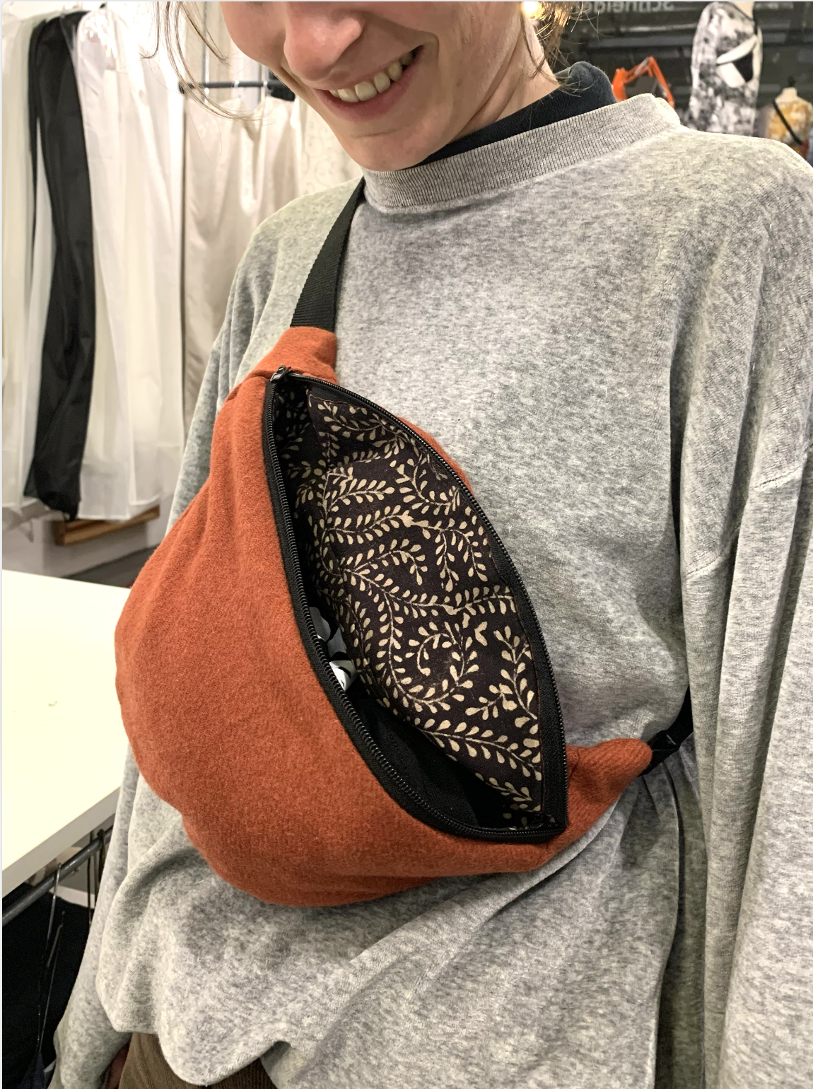 A woman wearing a gray long-sleeve shirt and an orange and black zippered bag with a patterned interior. She is smiling and in an indoor setting with clothing racks in the background.