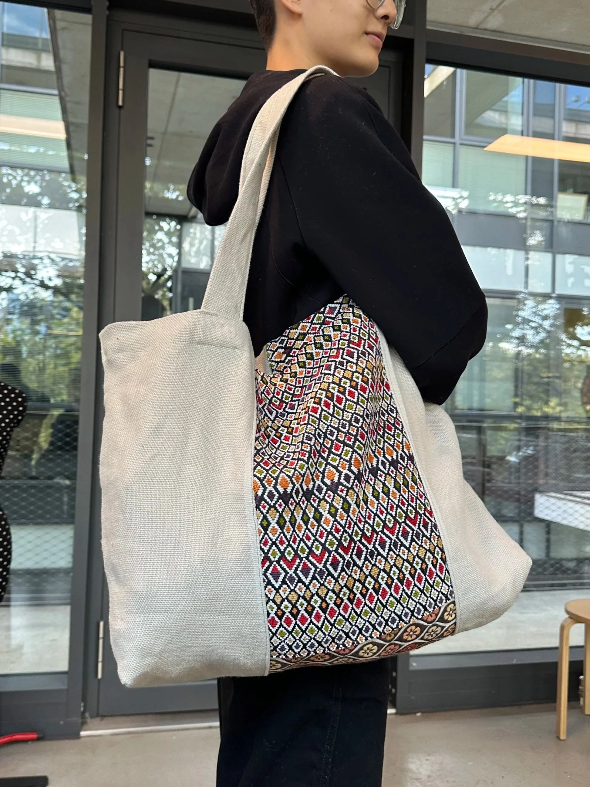 Person in black hoodie carrying a large cream tote bag with a multicolored geometric pattern on the side.