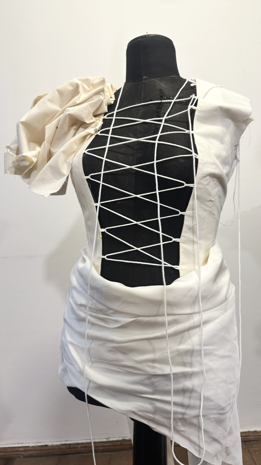 A dress form wearing an art piece with a corset-like black central fabric, white fabric panels, and white strings creating a crisscross pattern, with ruched white fabric on one shoulder.