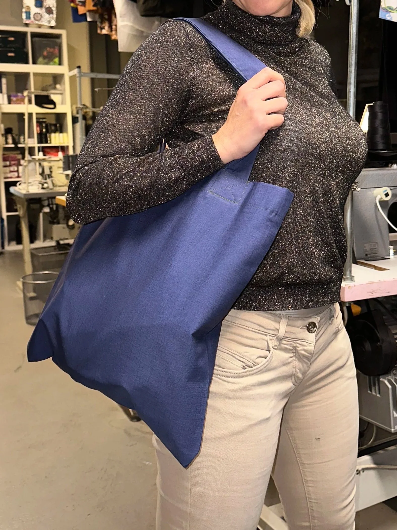 A woman holding a blue tote bag over her shoulder in a workspace or store with shelves and equipment in the background.