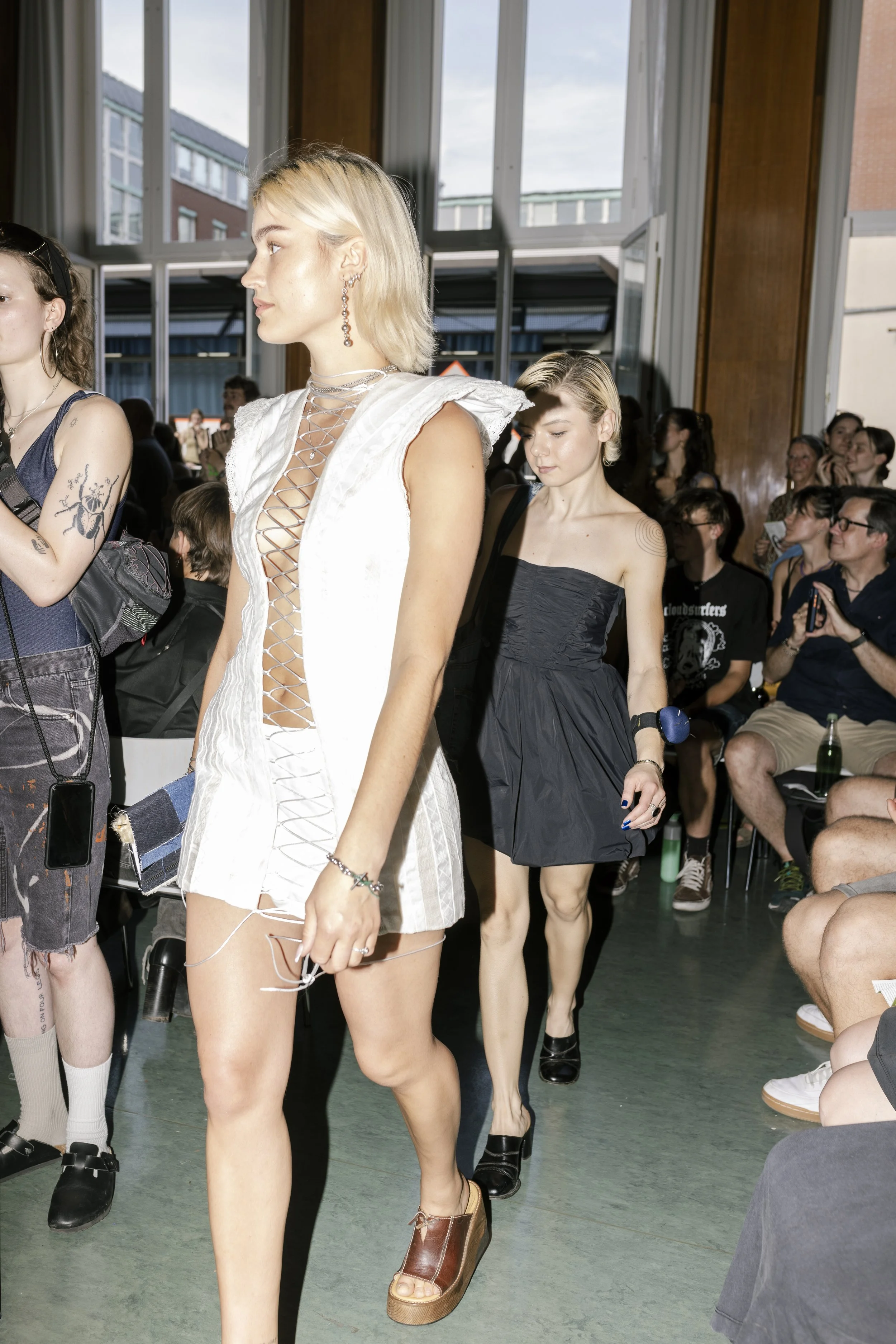 Models walking down the runway at a fashion show with audience seated on either side, in a well-lit indoor space with large windows.