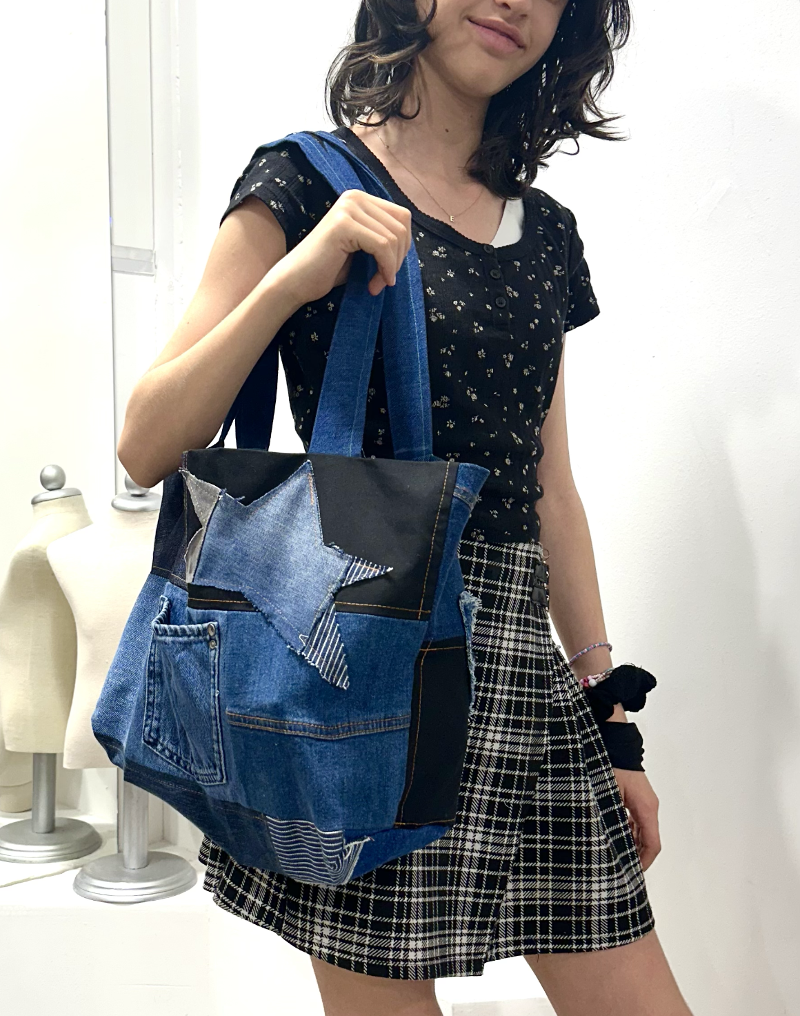 A woman is holding a denim tote bag with a star patch, wearing a black floral top and plaid skirt, standing against a white wall.