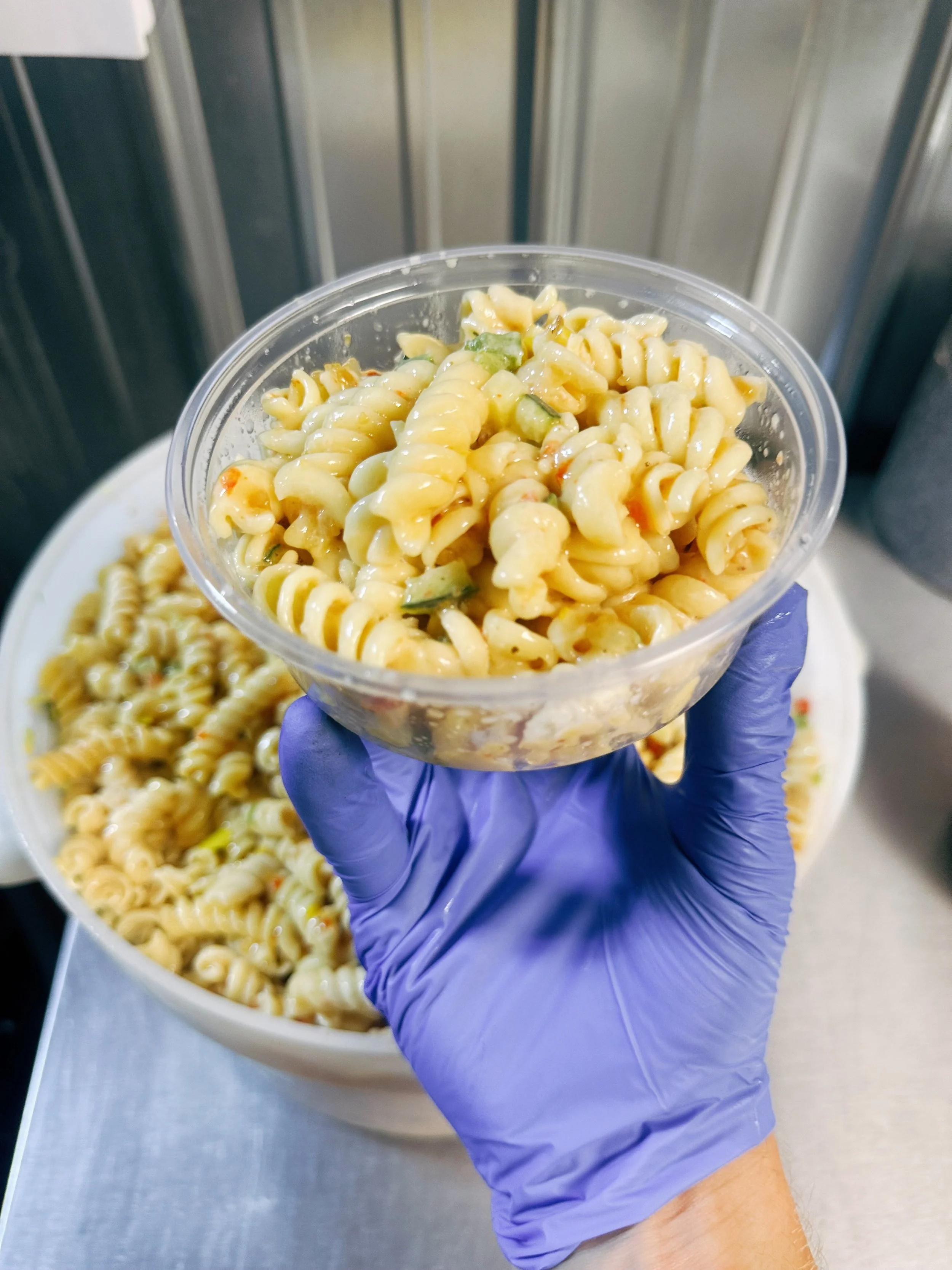 Person wearing a purple glove holding a plastic cup of rotini pasta salad with cucumber and dressing over a large bowl of similar salad.