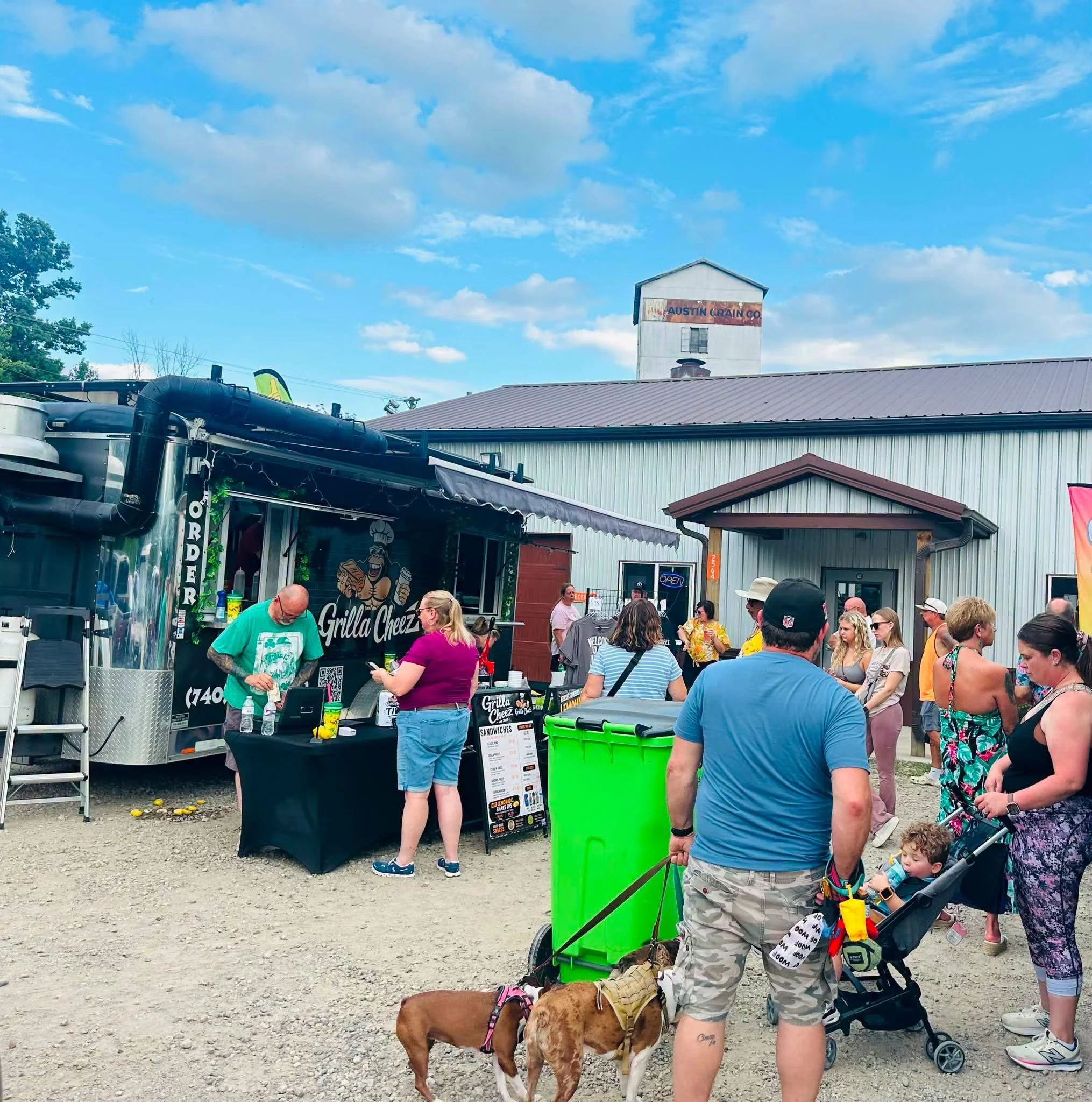 A group of people outdoors at what appears to be a food stand or festival, with some people holding dogs on leashes and others standing in line, under a partly cloudy sky.