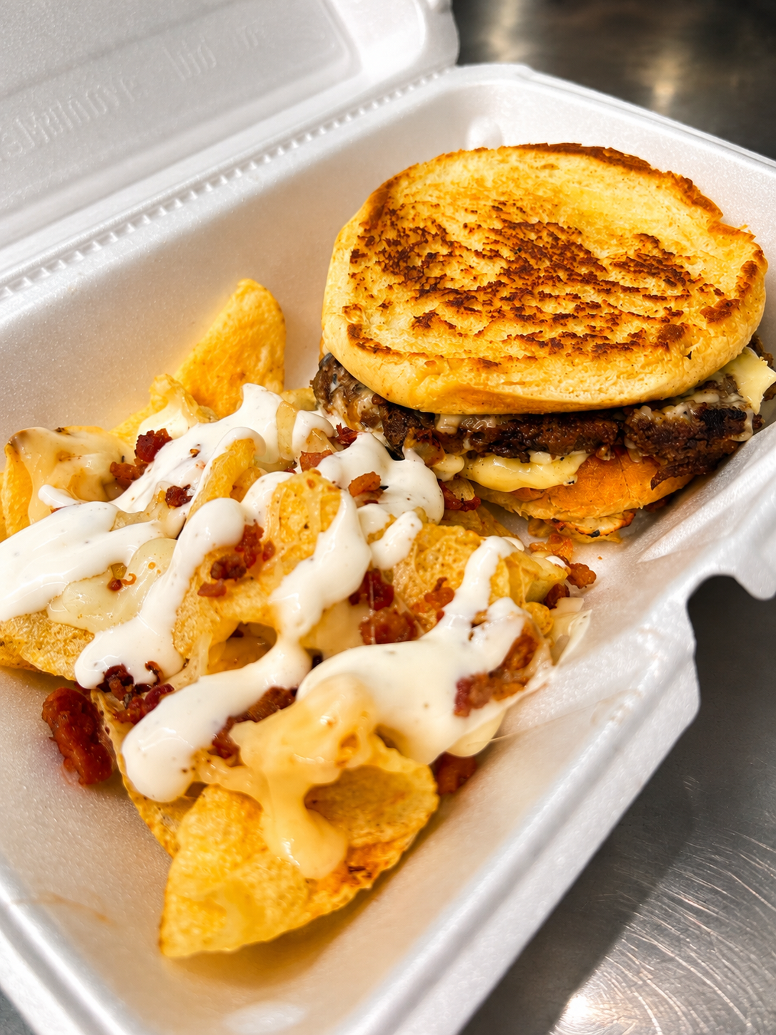 Cheeseburger with grilled bun and shredded cheese in a foam takeout container, next to loaded nachos topped with cheese, bacon bits, and sour cream.