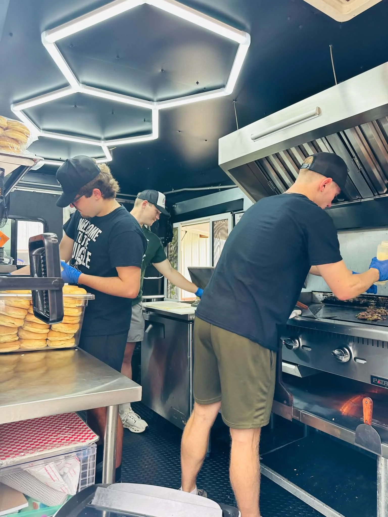 Three young men working in a food truck kitchen, preparing food on a stove with hamburgers and buns visible on a tray in the foreground.