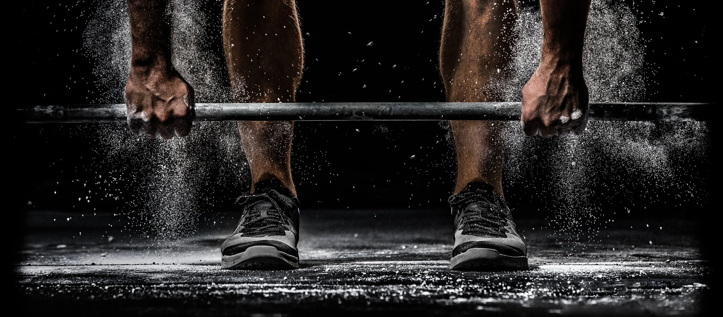 Person lifting a barbell with chalk-dusted hands and sneakers in a gym setting.
