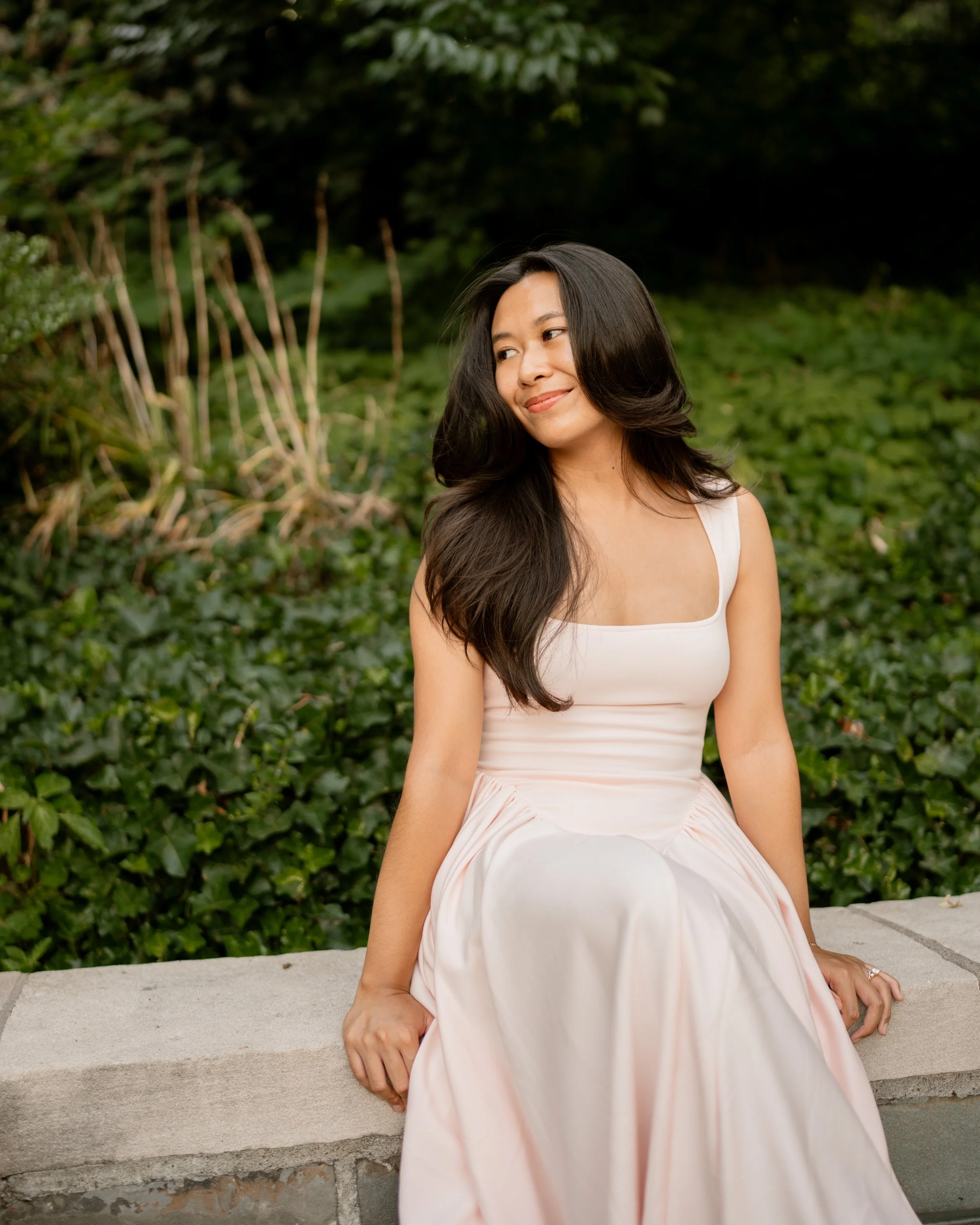 Calista Hennie, wearing a light pink dress and sitting in front of a green, nature background.