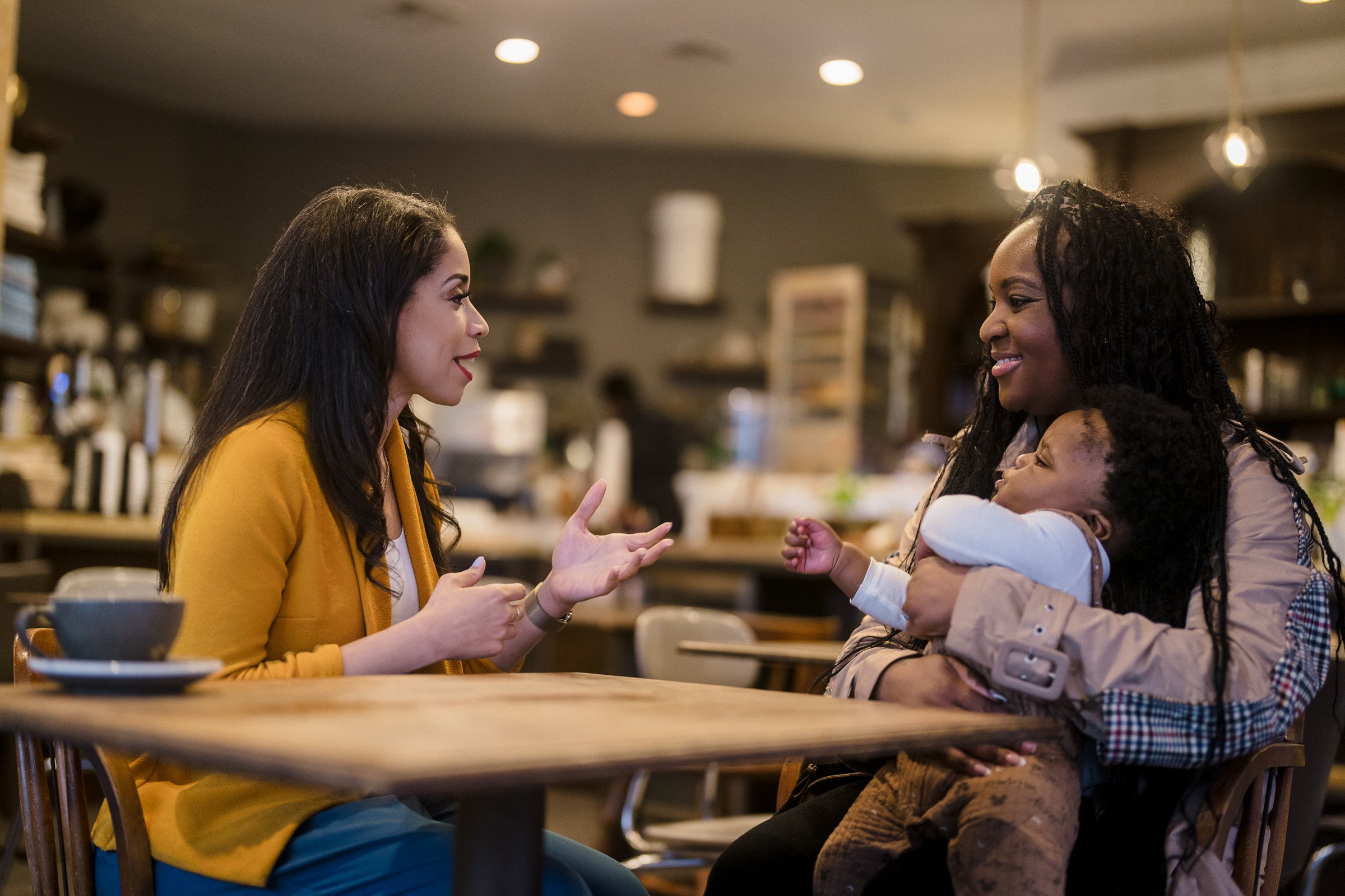 Two women sitting at a table in a café, one holding a baby.