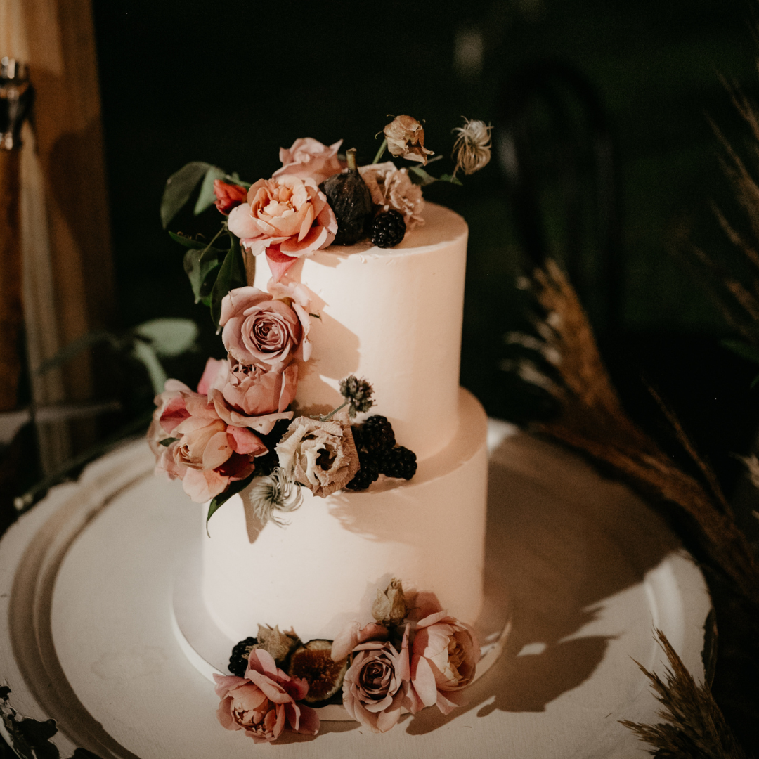 Two-tiered white cake decorated with pink roses and blackberries on a wooden table.