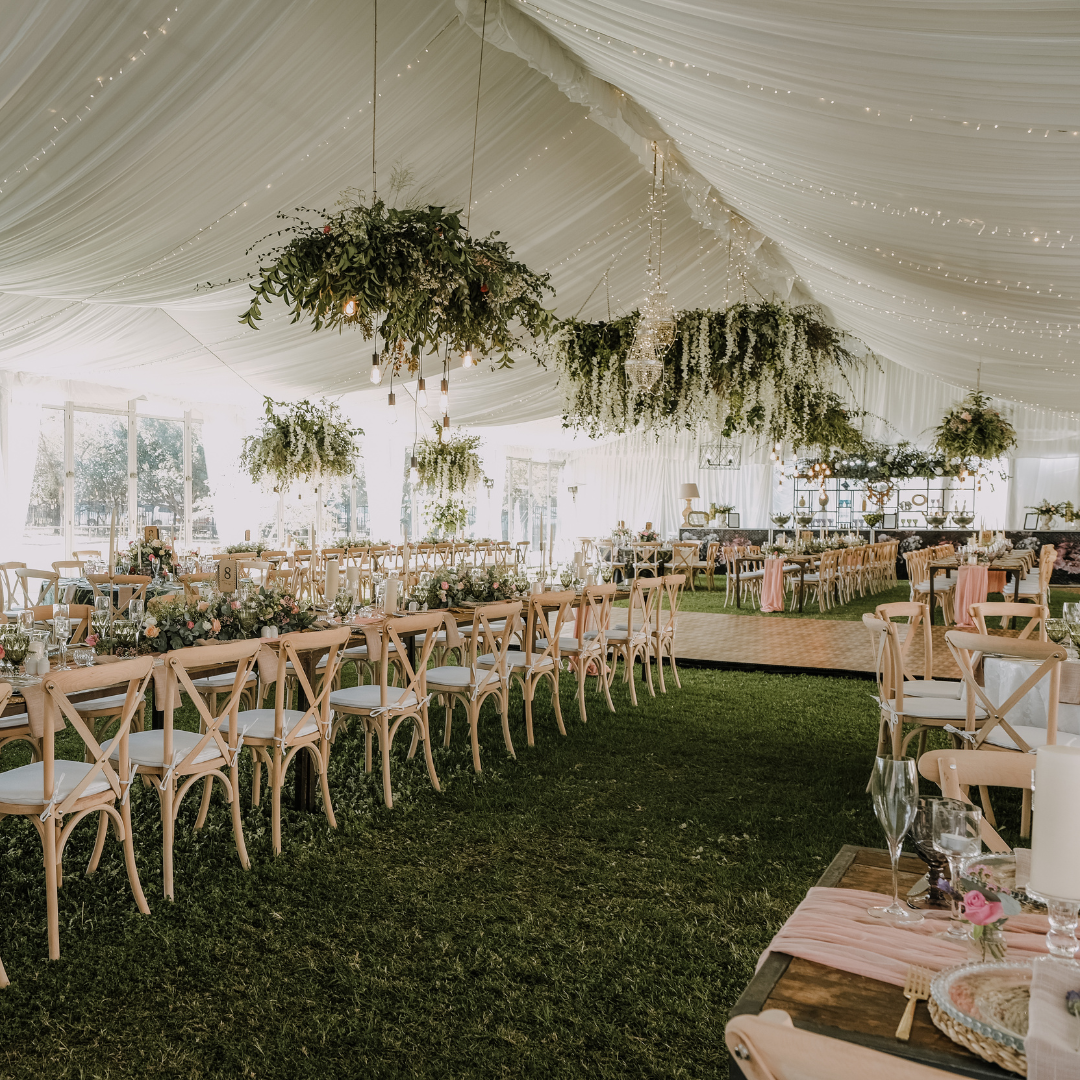 Elegant wedding reception in a tent with wooden chairs, decorated tables with floral arrangements, greenery chandeliers, and soft lighting.