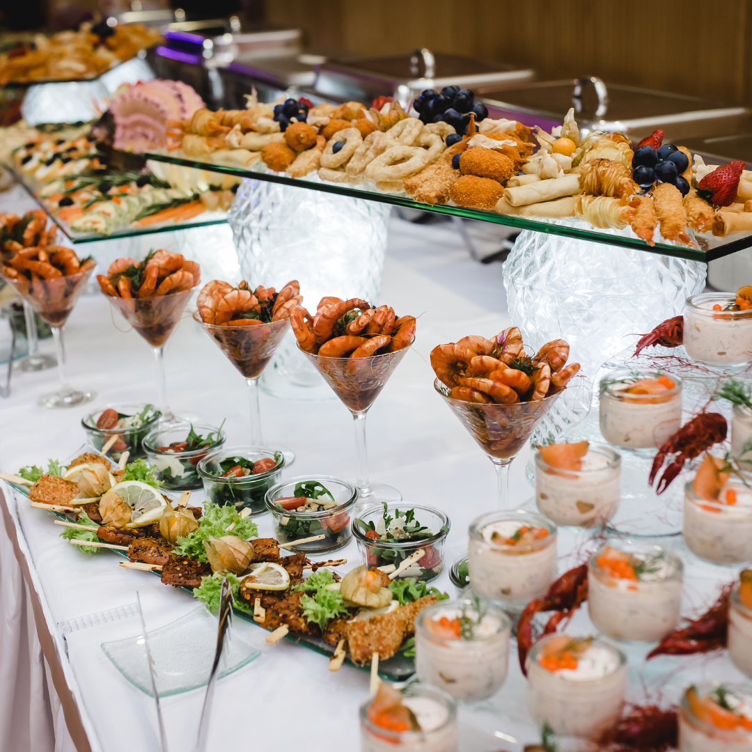 A buffet table with a variety of appetizers, including skewers, shrimp cocktail, fruit, and dessert cups.