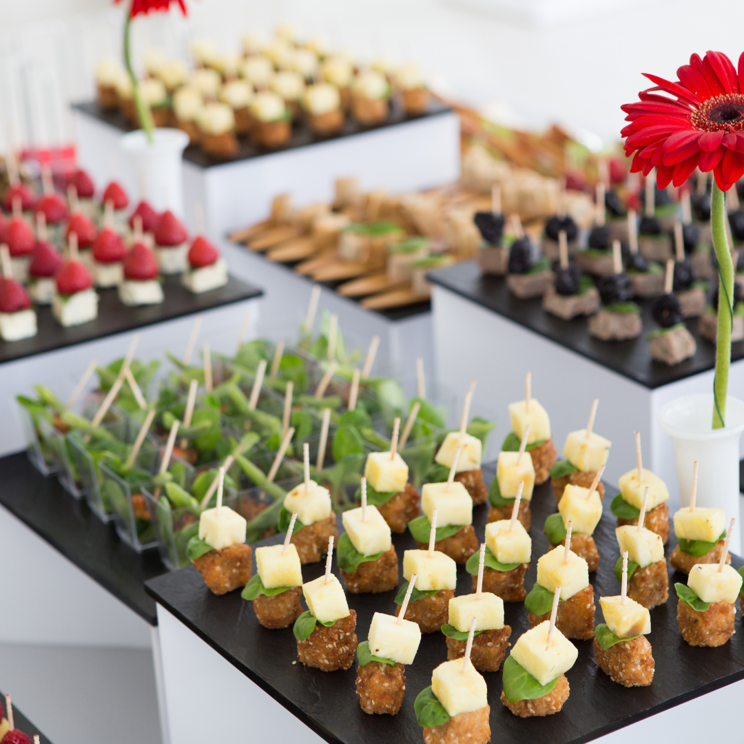 An array of assorted bite-sized appetizers, each skewered on toothpicks, including cheese cubes, fruit, and small savory bites, displayed on black slate serving platters with a red flower centerpiece.