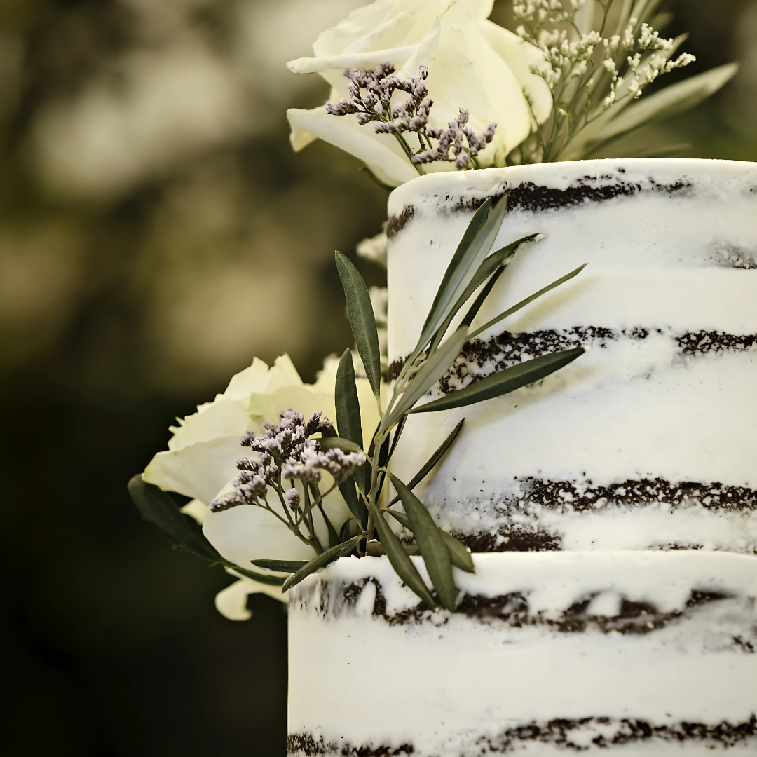 A two-tiered rustic naked cake with white frosting, accented with light-colored flowers and greenery, set against a blurred outdoor background.