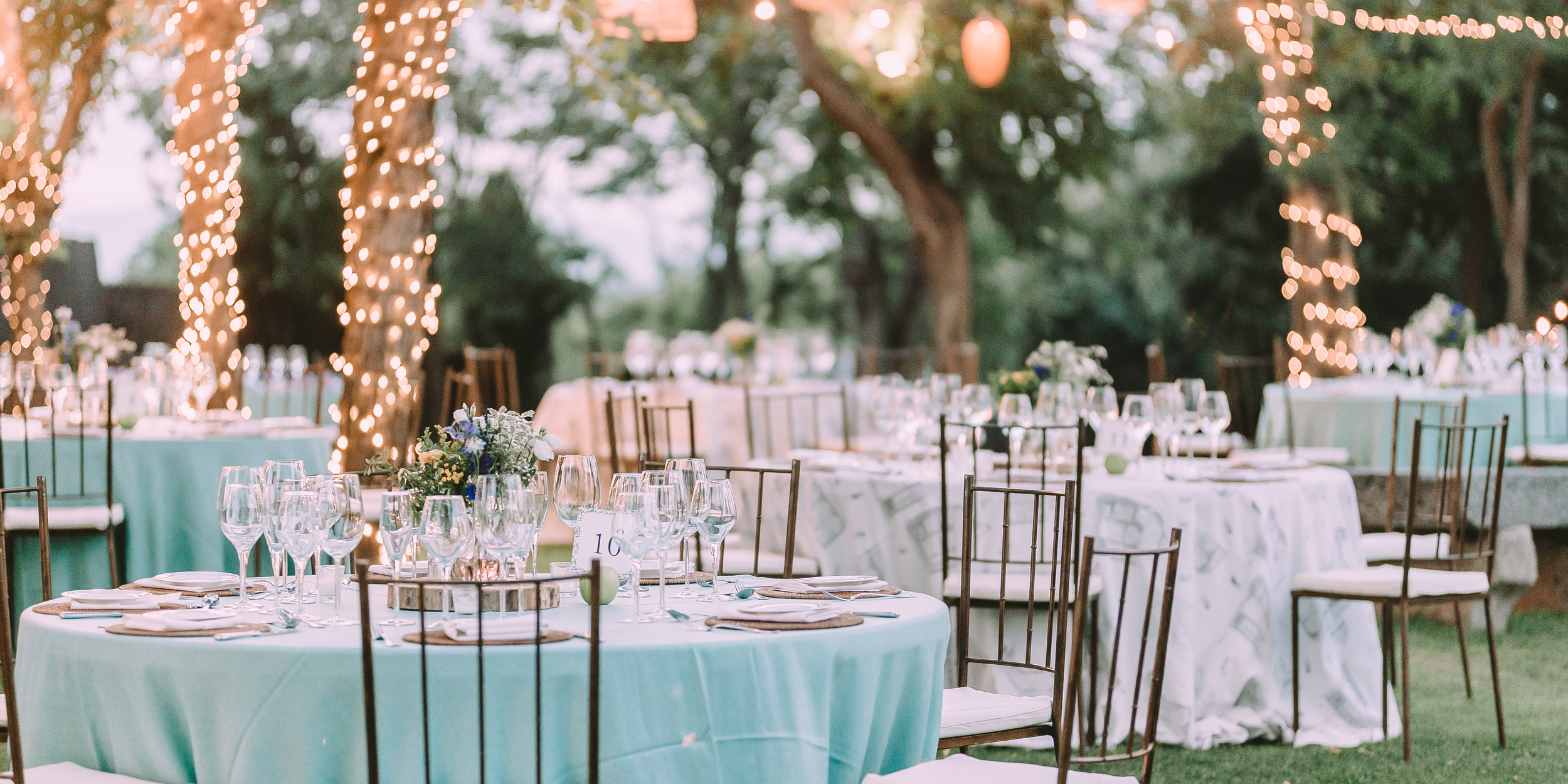 Outdoor wedding reception setup with round tables covered with white and turquoise tablecloths, surrounded by chairs and decorated with floral centerpieces. Trees wrapped in string lights provide a festive ambiance.