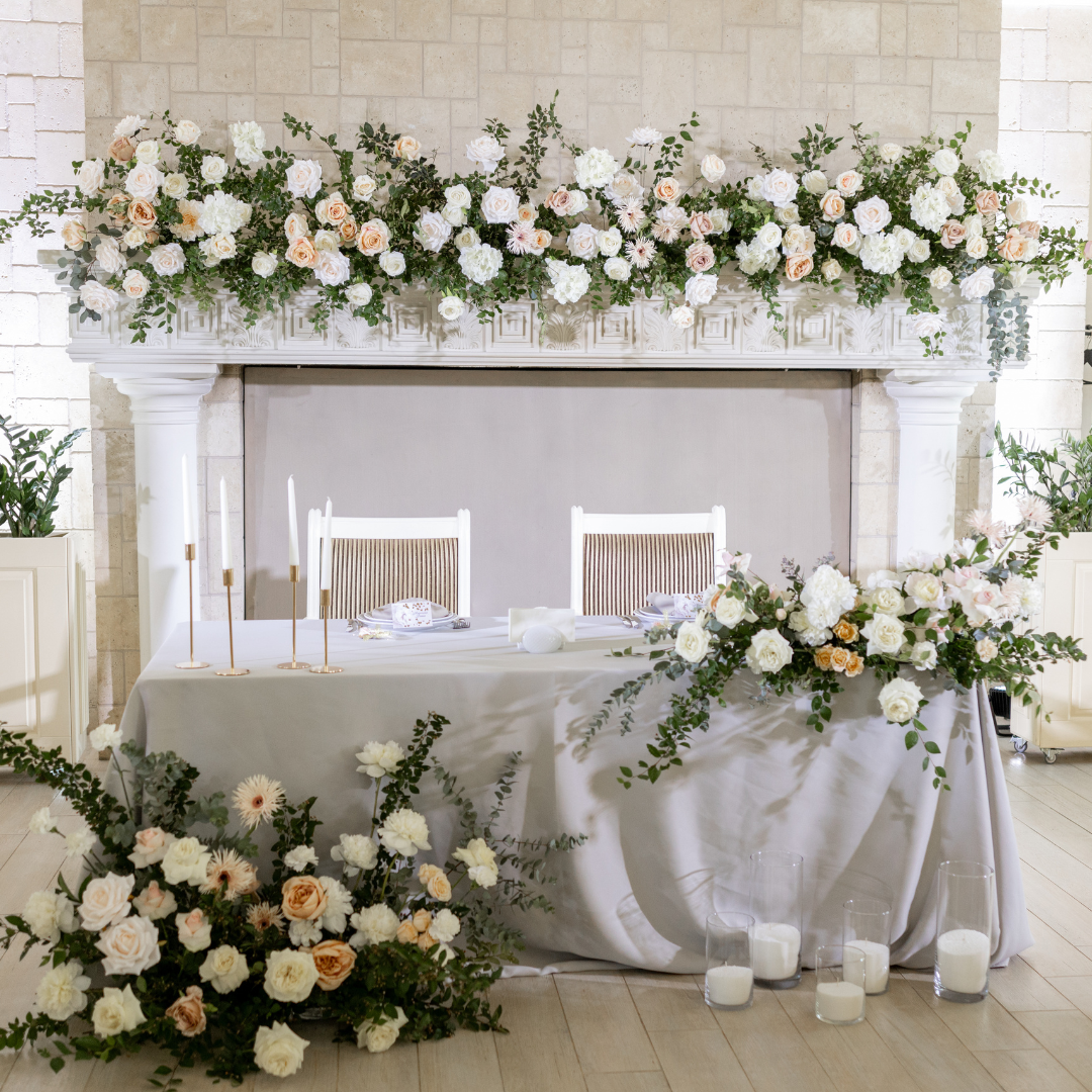Elegant wedding table setting with a floral arrangement backdrop, white chairs, tall candlesticks, and candles on the floor. The table is covered with a gray cloth and decorated with white and peach flowers.