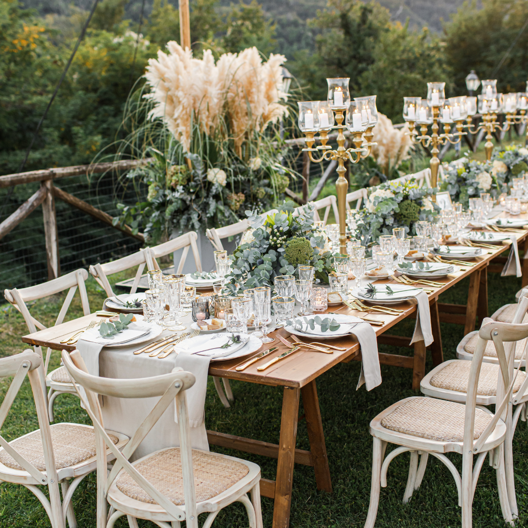 Elegant outdoor dining setup with a long wooden table, white chairs, floral centerpieces, and gold candlesticks, set on a lawn with greenery in the background.