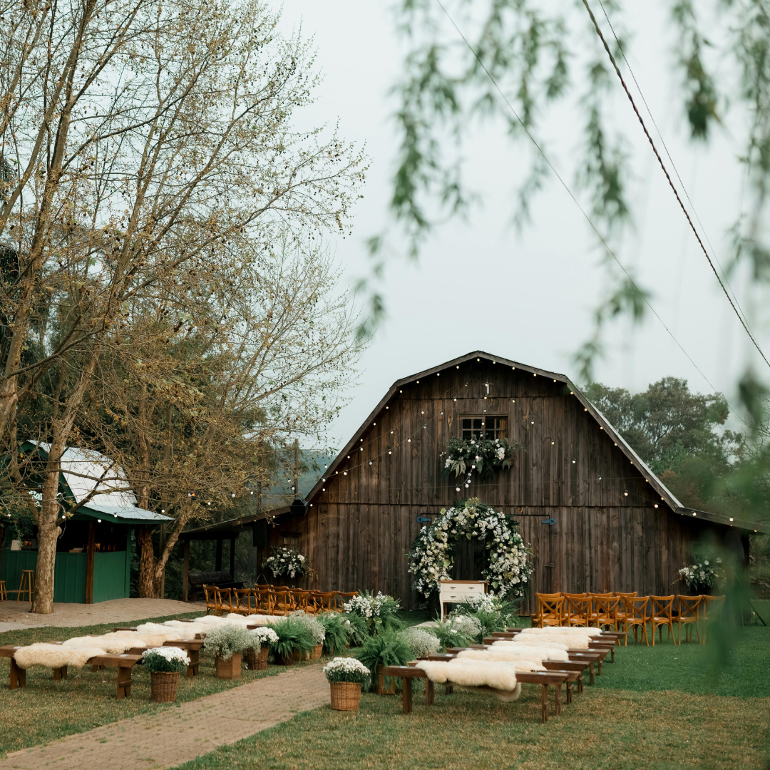 Rustic outdoor wedding venue with a decorated barn, floral arrangements, wooden benches with cushions, surrounded by trees and greenery.