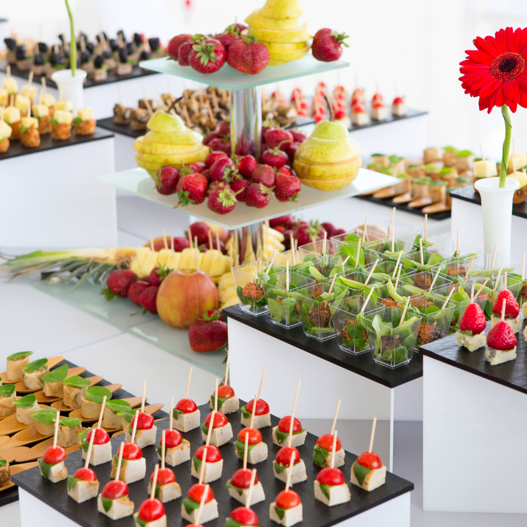 Assorted appetizers displayed on a white table, including fruit skewers with strawberries, pears, and pineapple, small appetizers with cherry tomatoes and cheese on sticks, leafy greens in clear cups, and a red flower as decoration.