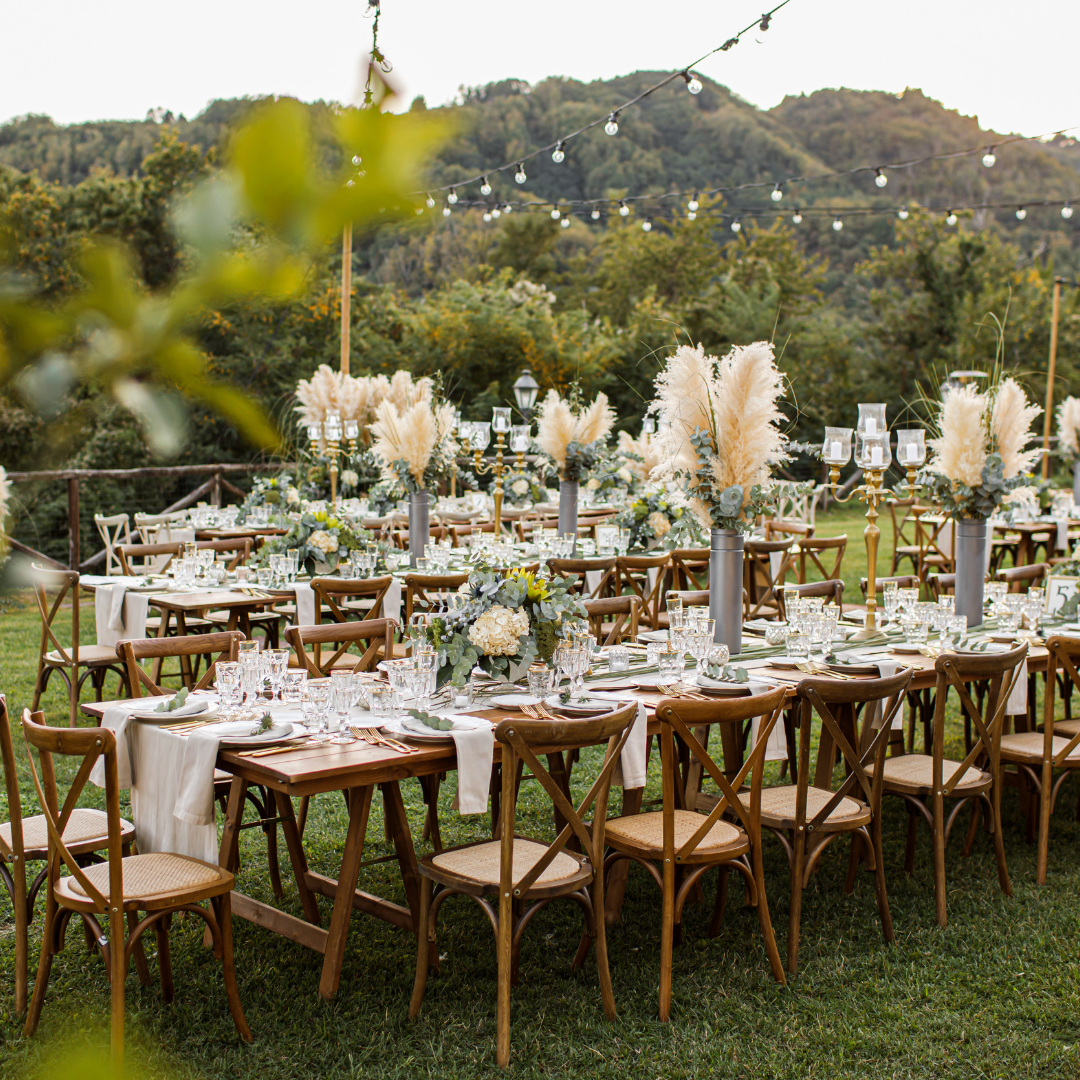 Outdoor wedding reception setup with wooden tables and chairs decorated with elegant centerpieces and string lights, surrounded by greenery and mountains.