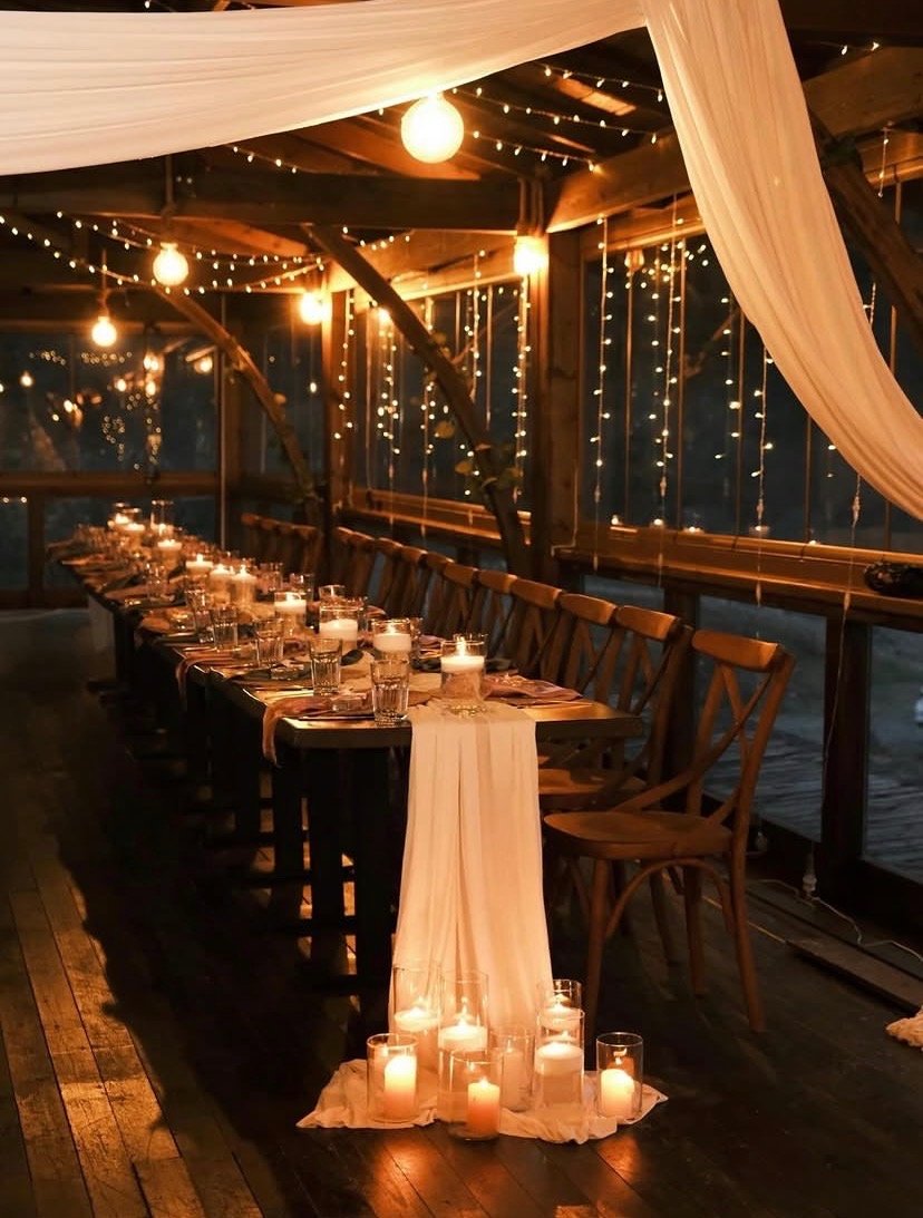 A warmly lit dinner setup with a long table, wooden chairs, candles, string lights, and fabric drapes, set in a rustic, enclosed space during the evening or night.
