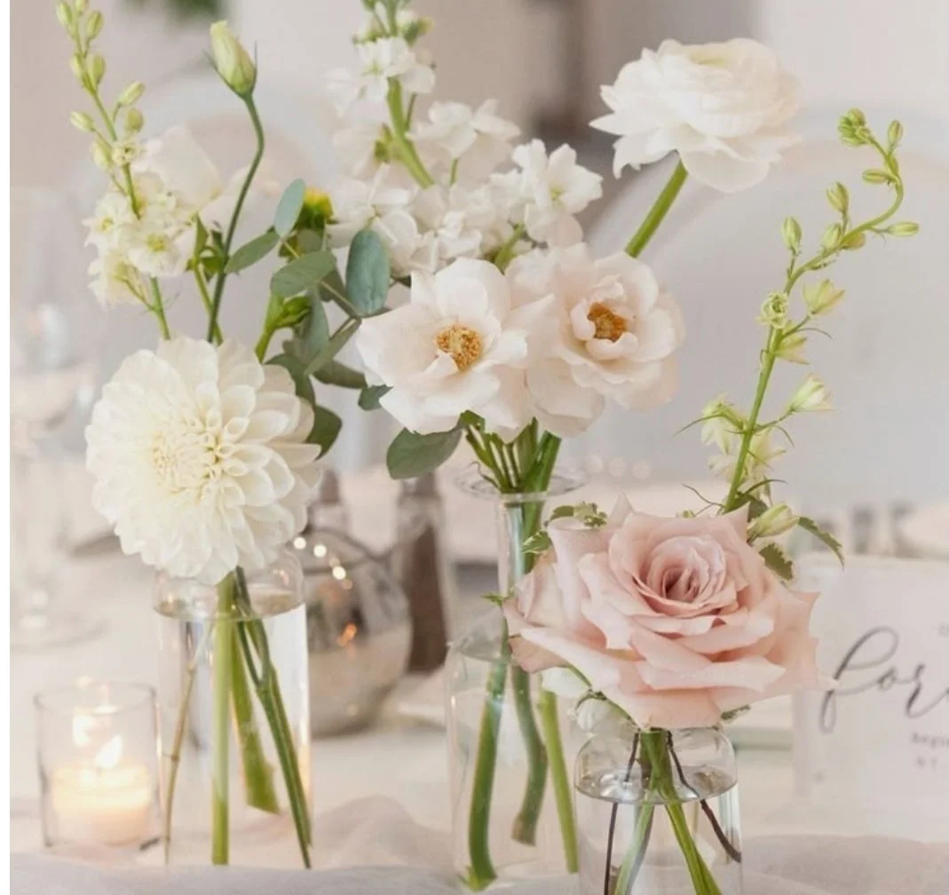 A collection of elegant white and blush pink flowers arranged in glass vases, set on a table with a lit candle nearby.
