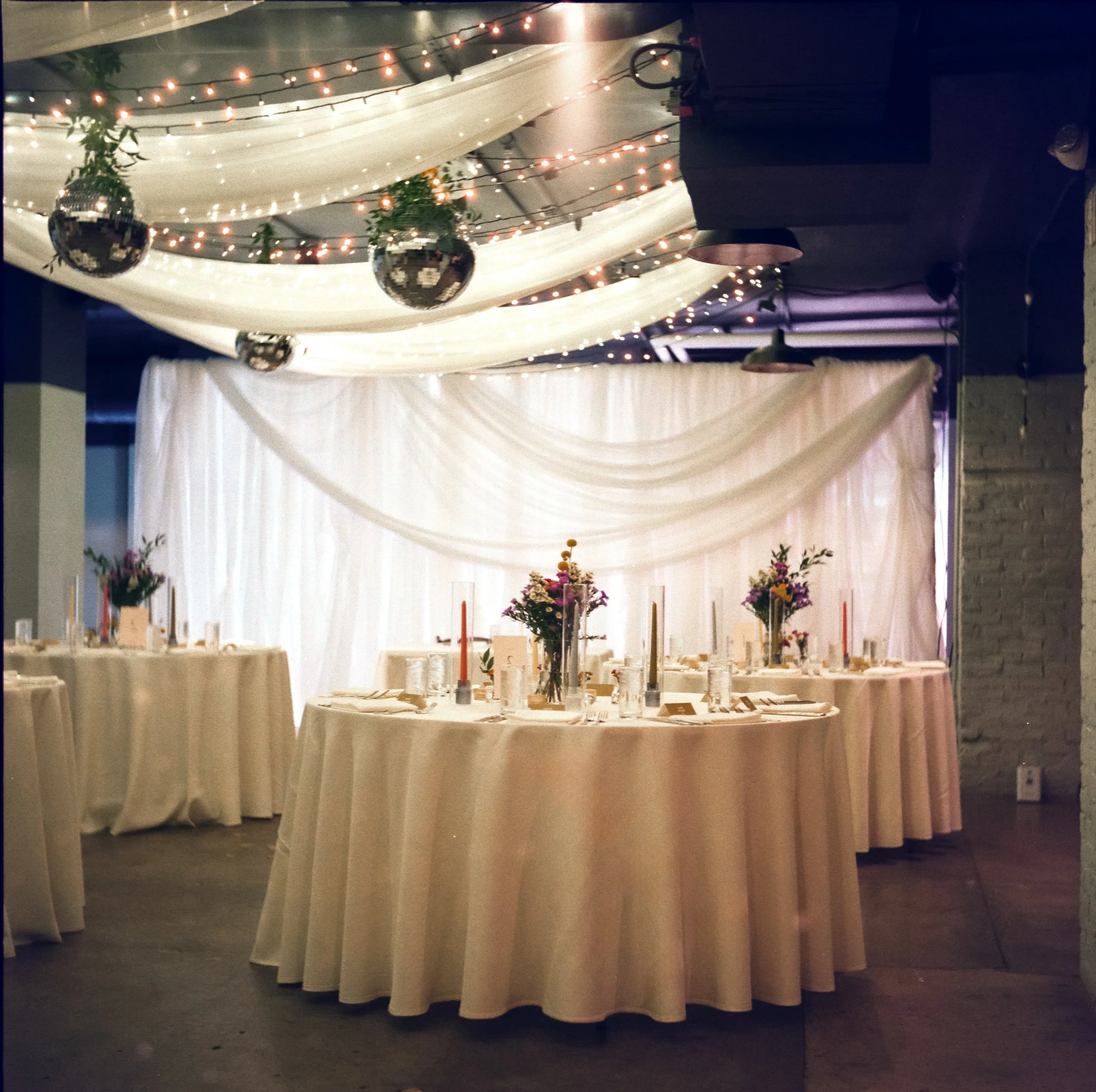 An event space with a long, round table covered with a white tablecloth, decorated with tall flower arrangements, candles, and place settings. The backdrop features draped white fabric, string lights, and hanging greenery. Disco balls hang from the ceiling, and the overall lighting is soft and warm.