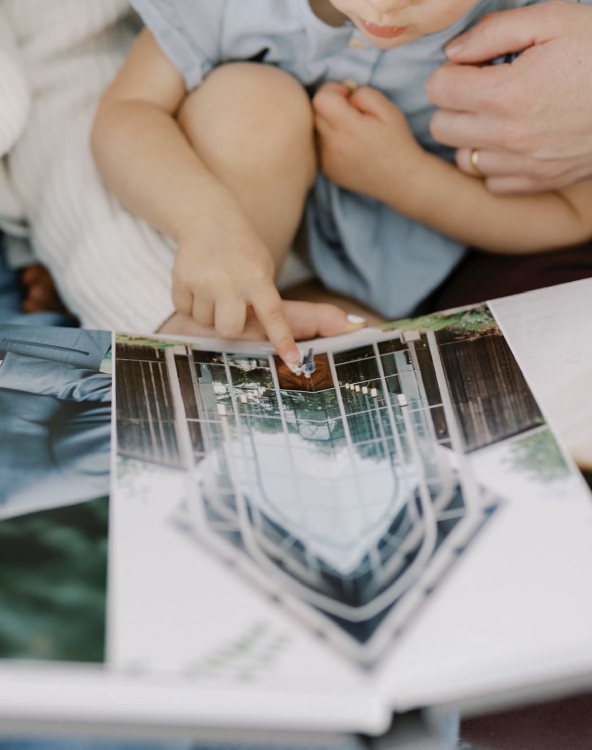 Child pointing at a photo in an open book, with an adult's hand gently touching the child.