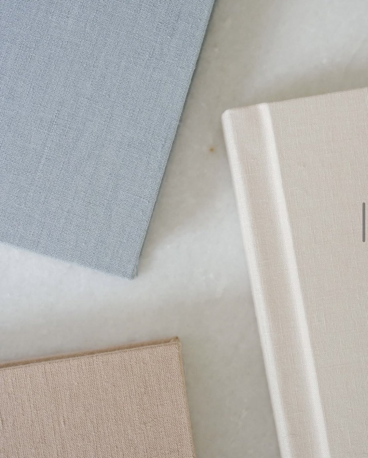 Flat lay of three linen-covered books in light gray, cream, and beige on a marble surface.