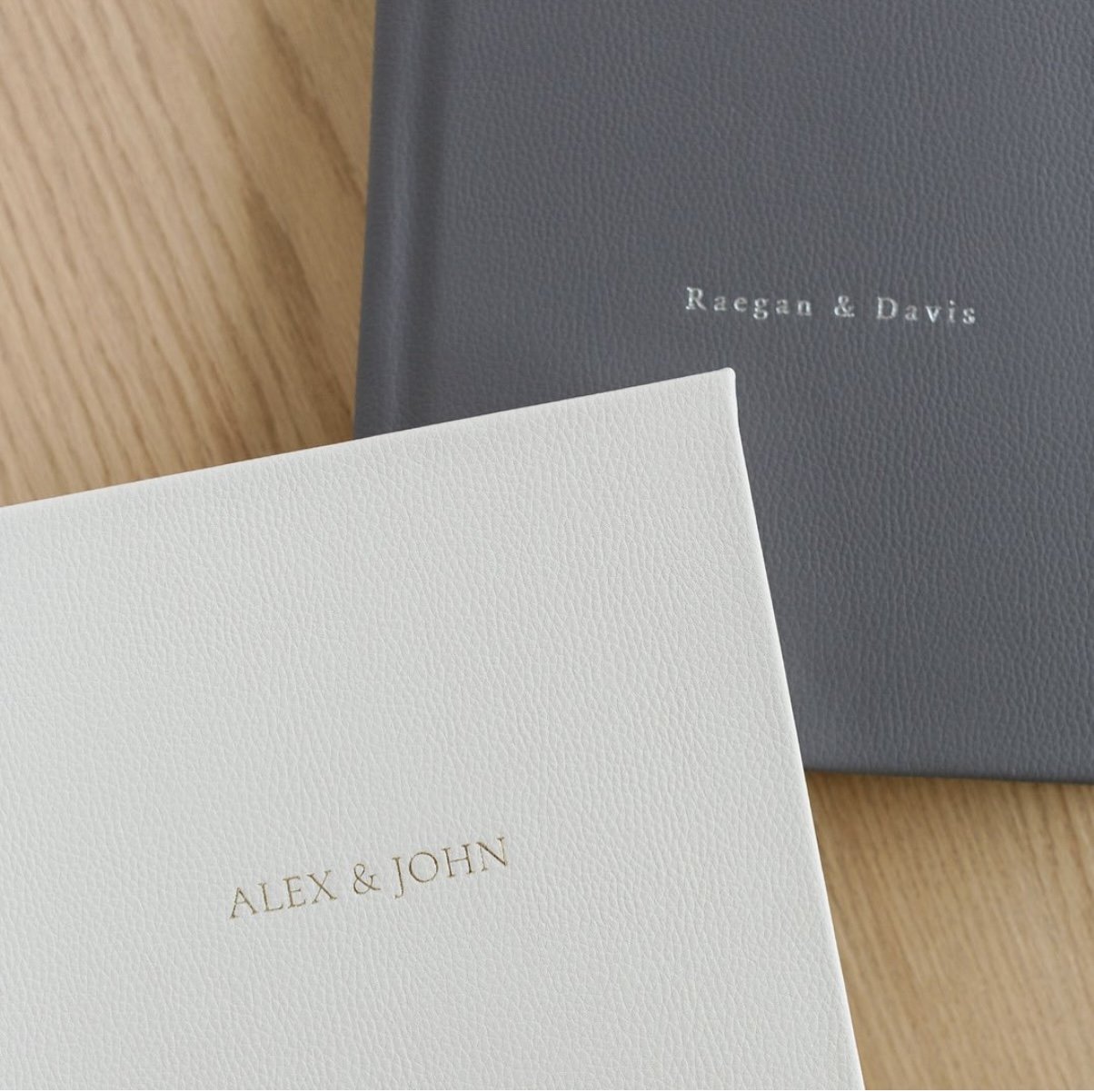 Two leather-bound books with embossed names on the covers, "Alex & John" on a white book and "Raegan & Davis" on a gray book.