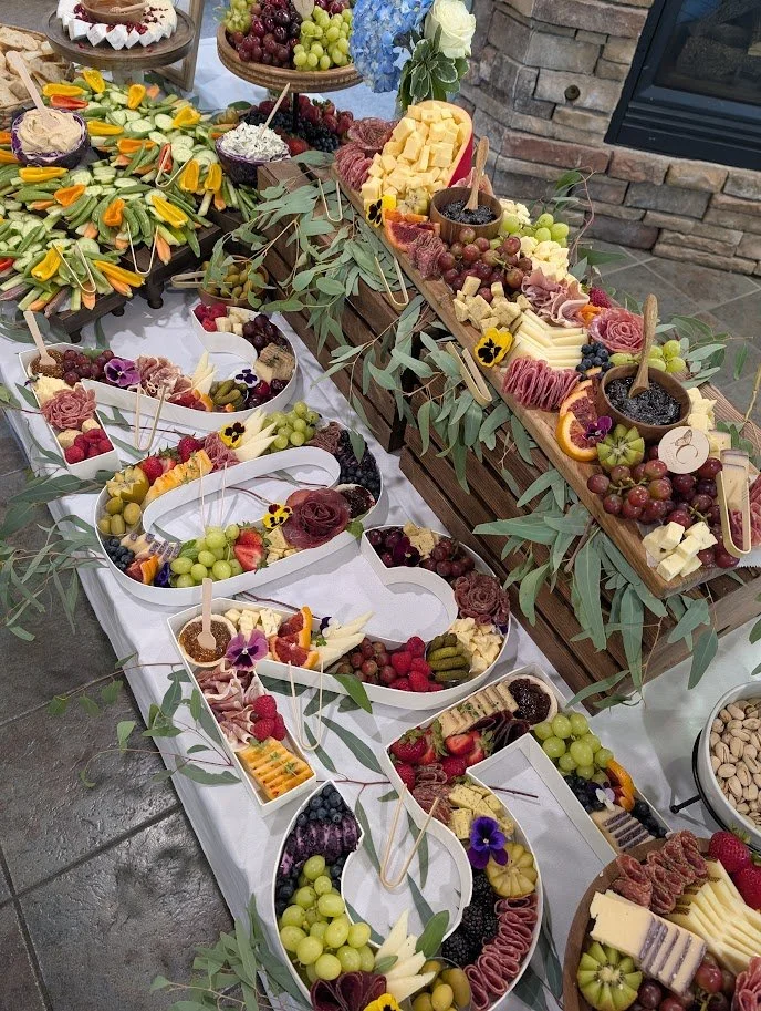 A table decorated with a cheese and fruit platter display, featuring grapes, berries, cheese blocks, and floral accents.
