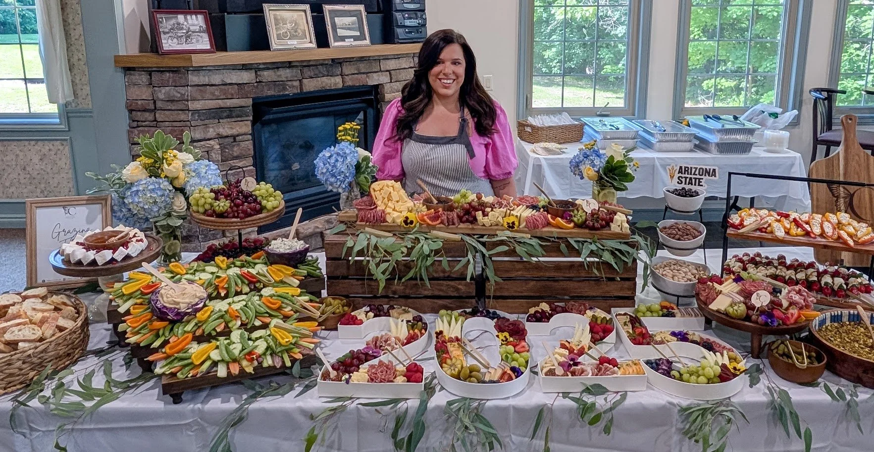A woman standing behind a table filled with various fruits, cheese, and vegetable platters at a buffet-style event. The room has large windows, a stone fireplace, and framed photos on the mantle. There are flowers and greenery decorating the table.