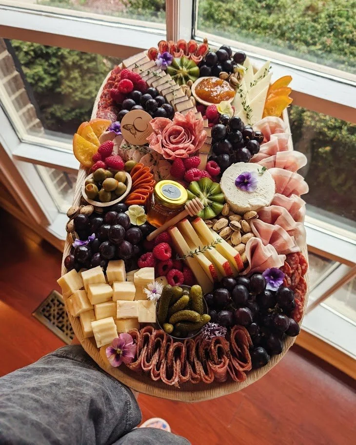 A large charcuterie board with assorted cheeses, grapes, berries, honey, crackers, and garnishes, placed on a wooden surface near a window.