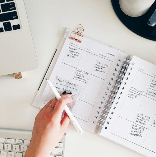Open planner with handwritten notes and appointments on a white desk, near a computer keyboard and a laptop.