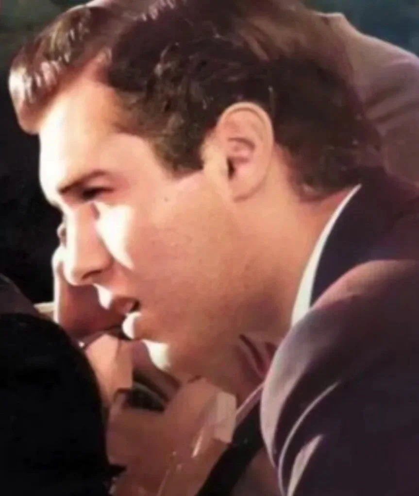 Close-up of a man with brown hair talking to a woman with dark hair, both dressed in formal attire, in a dimly lit setting.