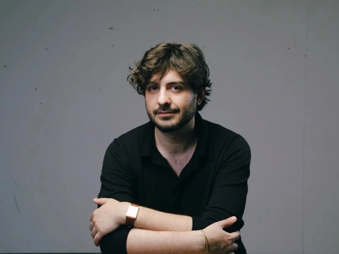 Un uomo con capelli ricci e occhiali indossa una maglietta nera e si trova di fronte a uno sfondo grigio. La persona ha un aspetto rilassato e sorride leggermente.