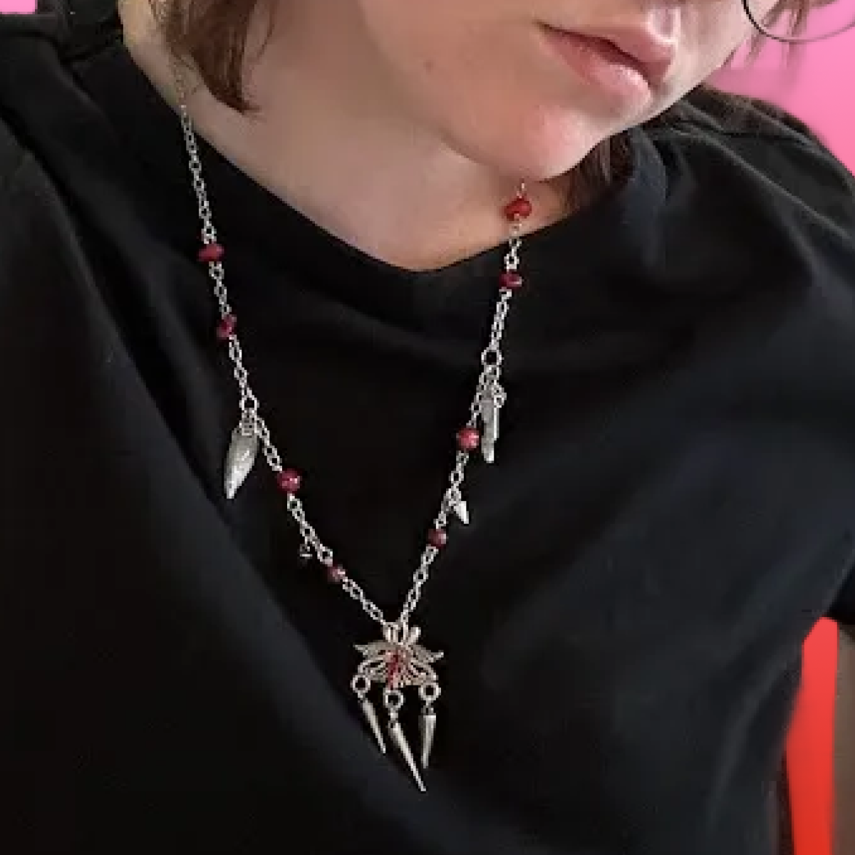 A person wearing a black top and two layered silver necklaces with red beads and pendant charms, including a winged owl and dagger design.