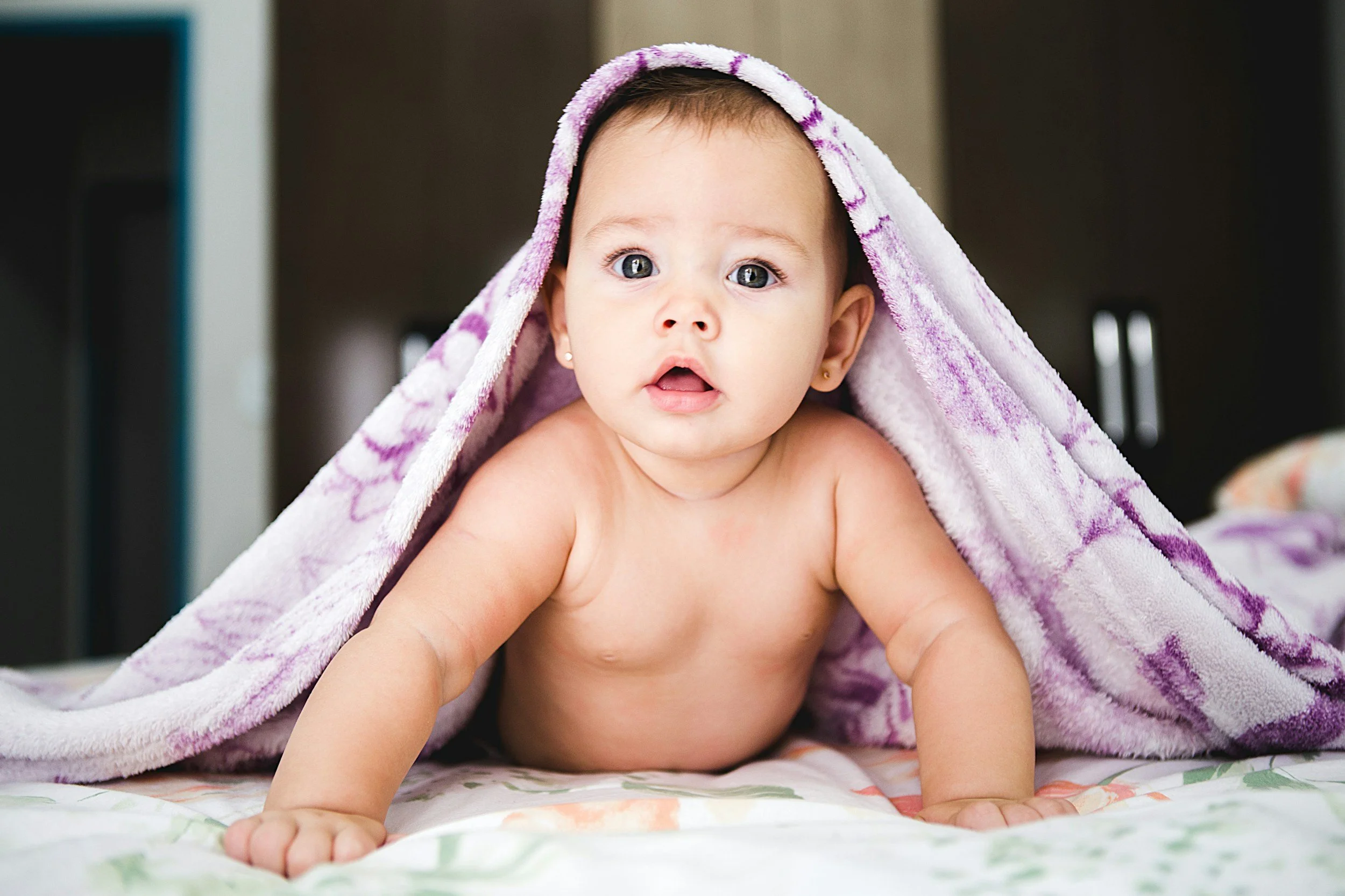 A baby with blue eyes, lying on their stomach on a bed, with a purple and white towel draped over their head.