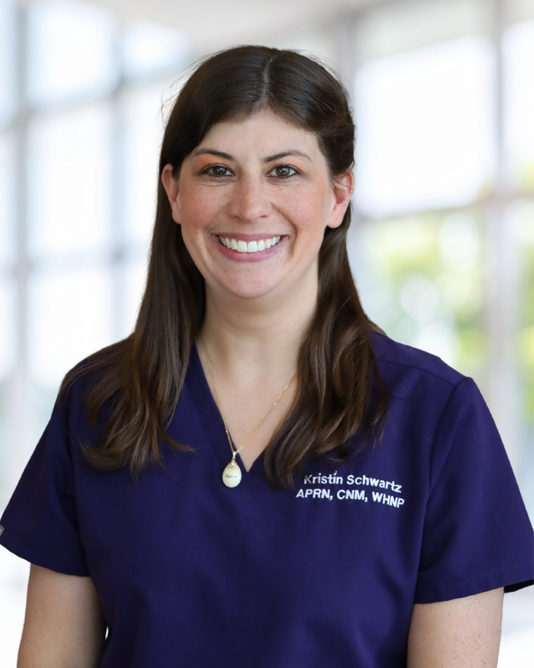 A woman smiling, wearing a navy blue medical scrub top with the embroidered text 'Kristin Schwartz APRN, CNM, WHNP,' standing in front of a bright, windowed background.