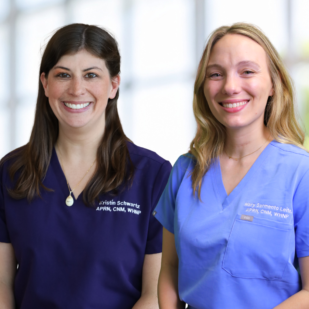 Two women wearing medical scrubs, smiling, standing indoors with a bright background.