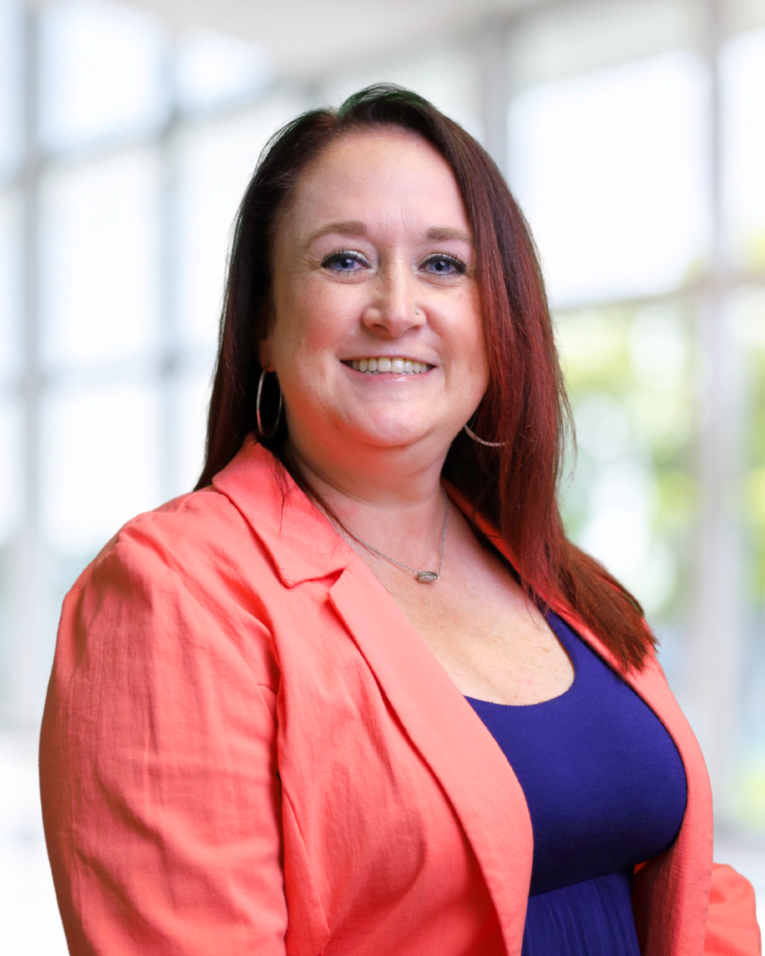 A smiling woman with reddish-brown hair, wearing a coral blazer over a navy blue top, standing in a bright indoor setting with large windows and blurred greenery in the background.