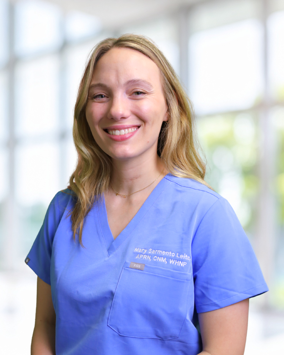 Portrait of a smiling female nurse or healthcare worker wearing blue scrubs in a well-lit hospital or clinic setting.