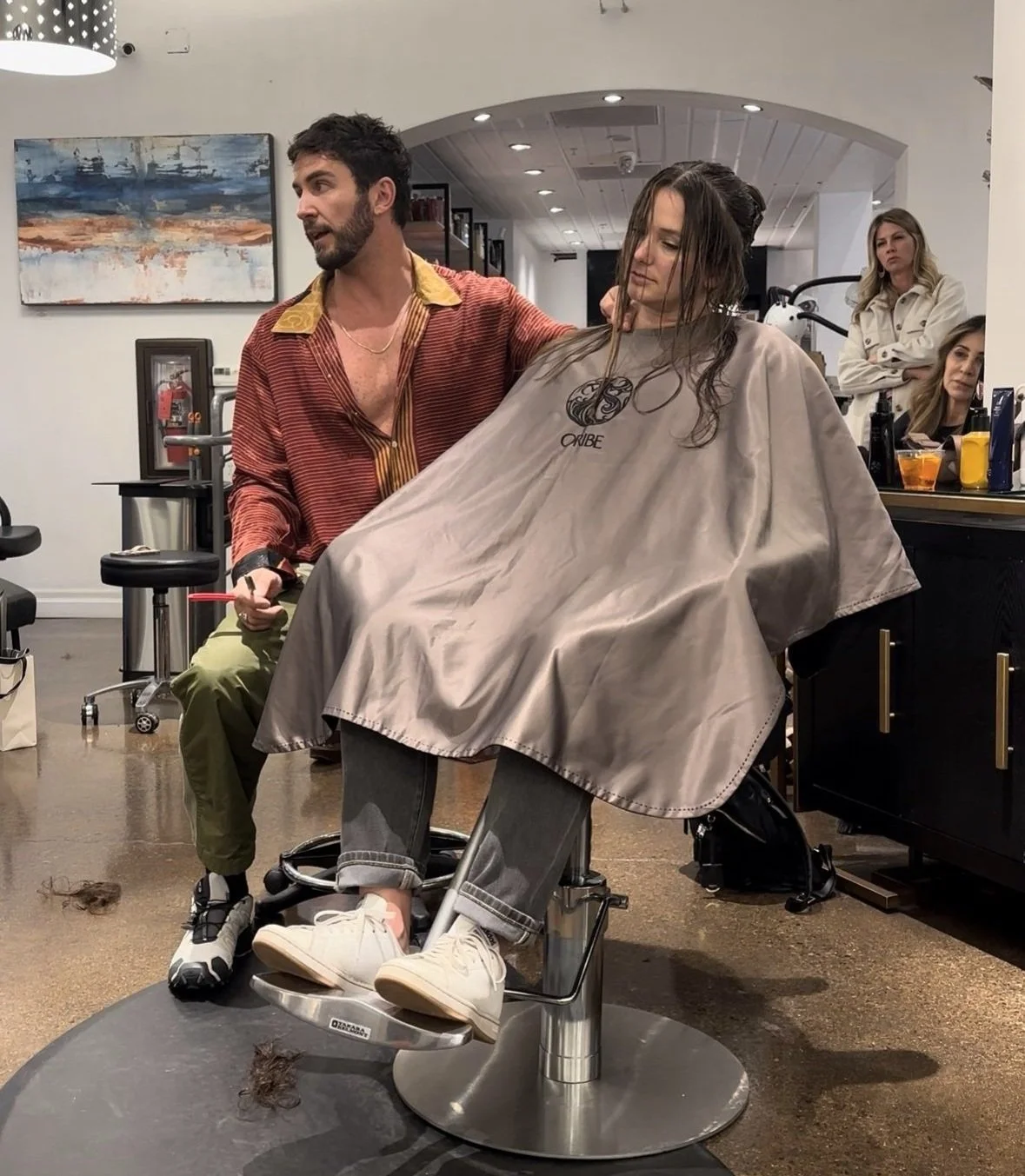 Hair salon scene with stylist and client getting a haircut under a cape, surrounded by spectators and salon equipment.