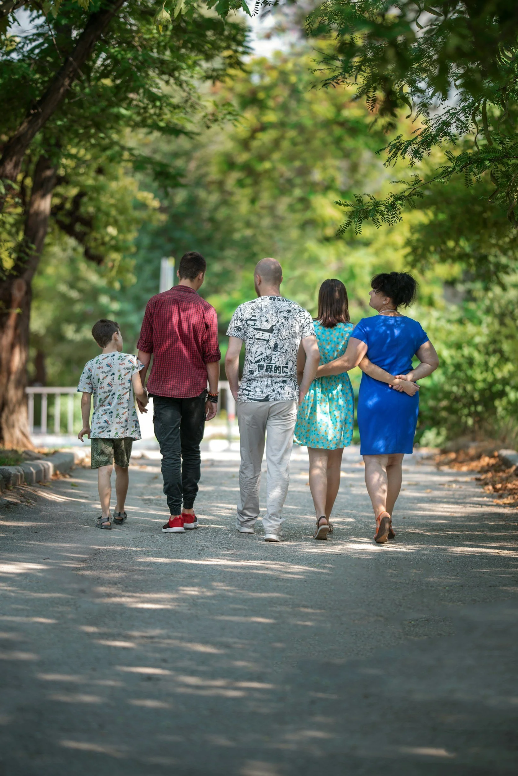 A group of five people walking on a tree-lined pathway, viewed from behind, in a park or wooded area.